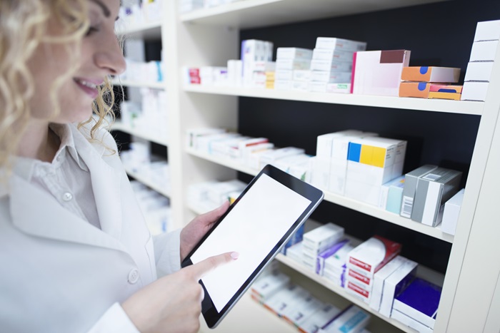 Pharmacist holding tablet by the shelf full of medicine in drug store and checking medication availability and specification.